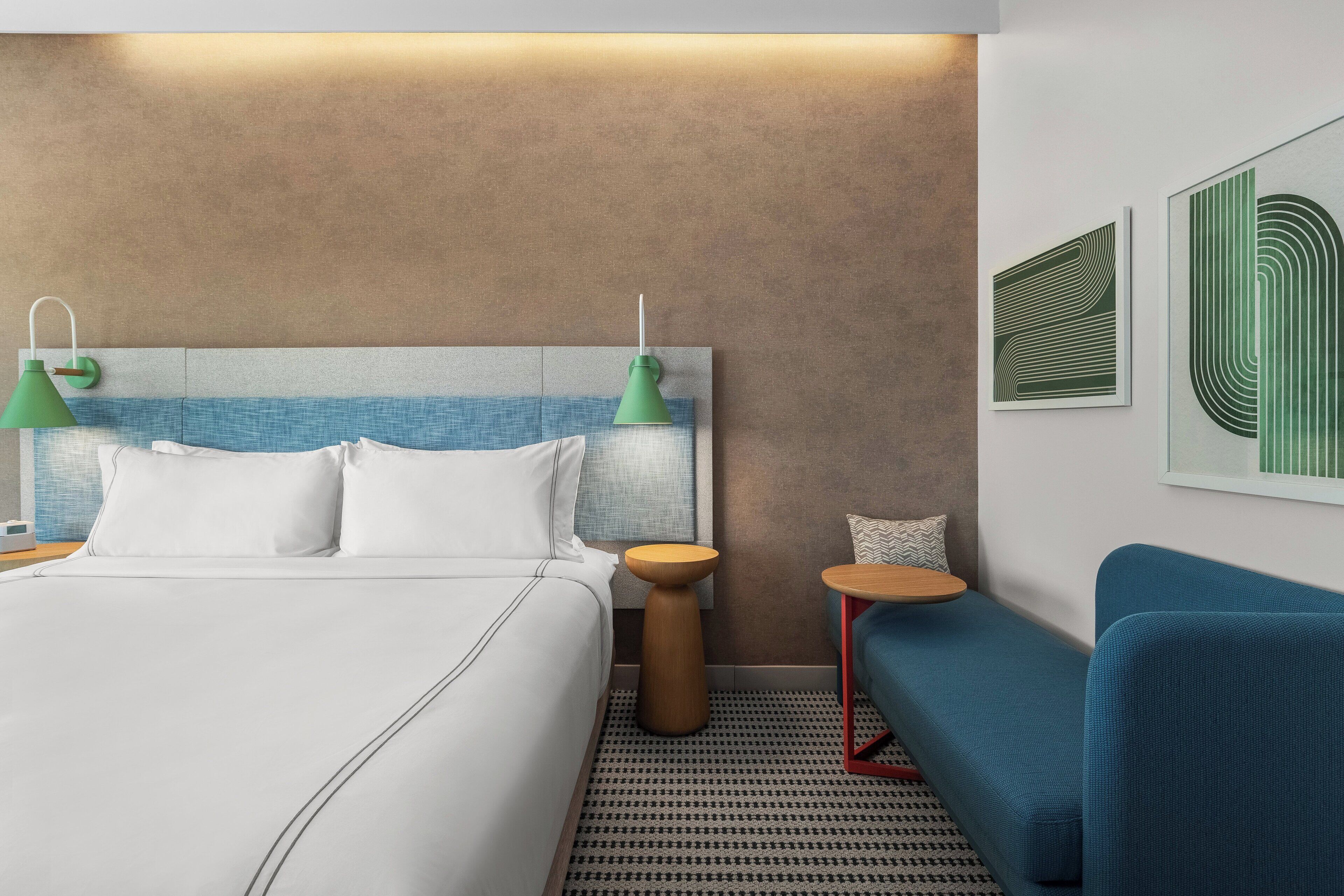 Een moderne hotelkamer met een groot bed, twee nachtkastjes, een blauwe bank en kunstwerken aan de muur.