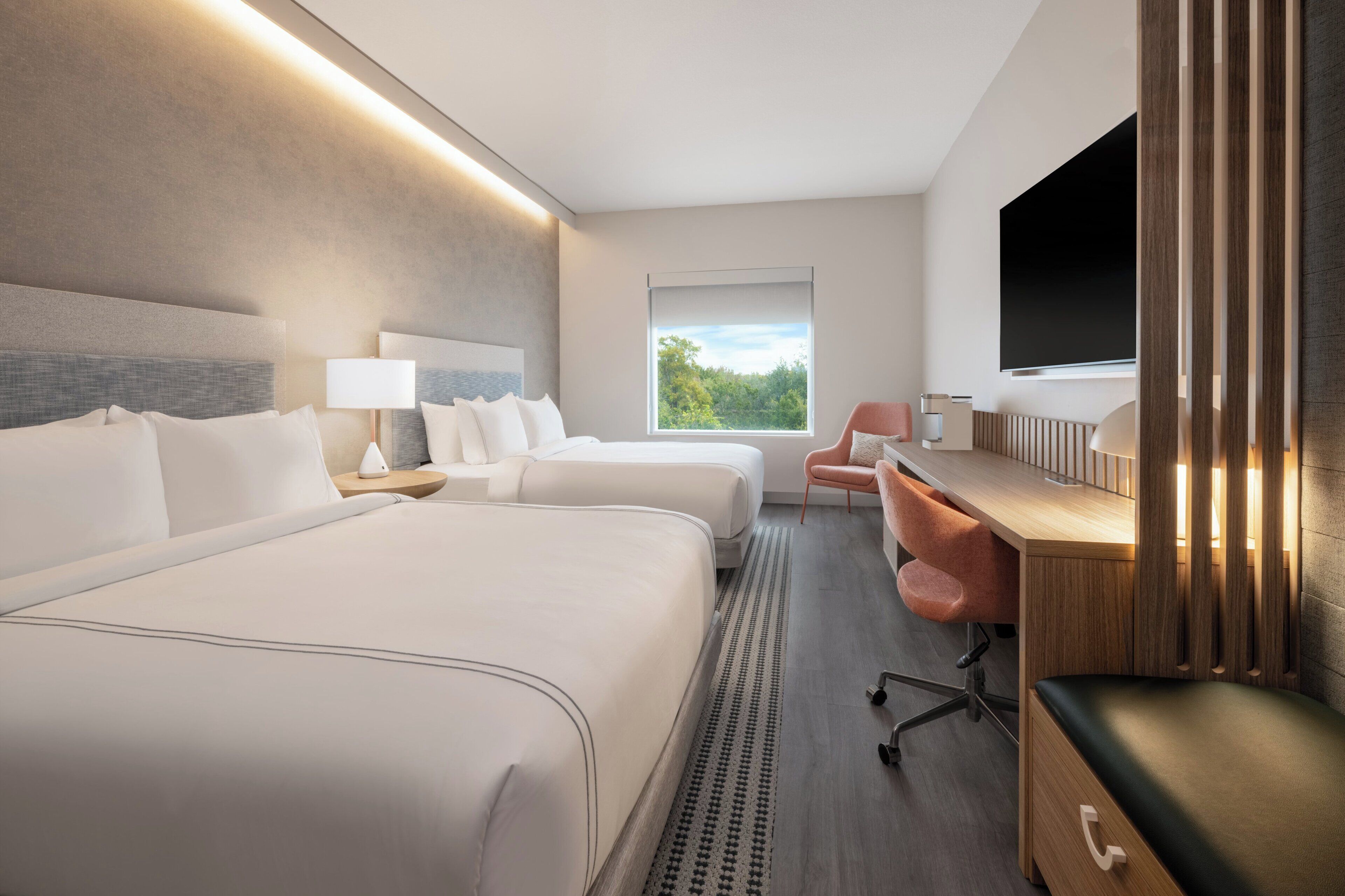 Habitación de hotel con dos camas, un escritorio, una silla, un televisor y una ventana con vista a árboles.