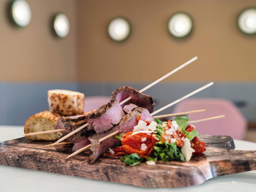 Een houten plank met spiezen vol vlees, een stuk brood en een salade.
