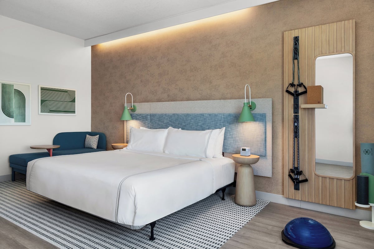 Una habitación de hotel moderna con una cama grande, dos mesitas de noche, un televisor montado en la pared, un espejo y una esterilla de yoga en el suelo.