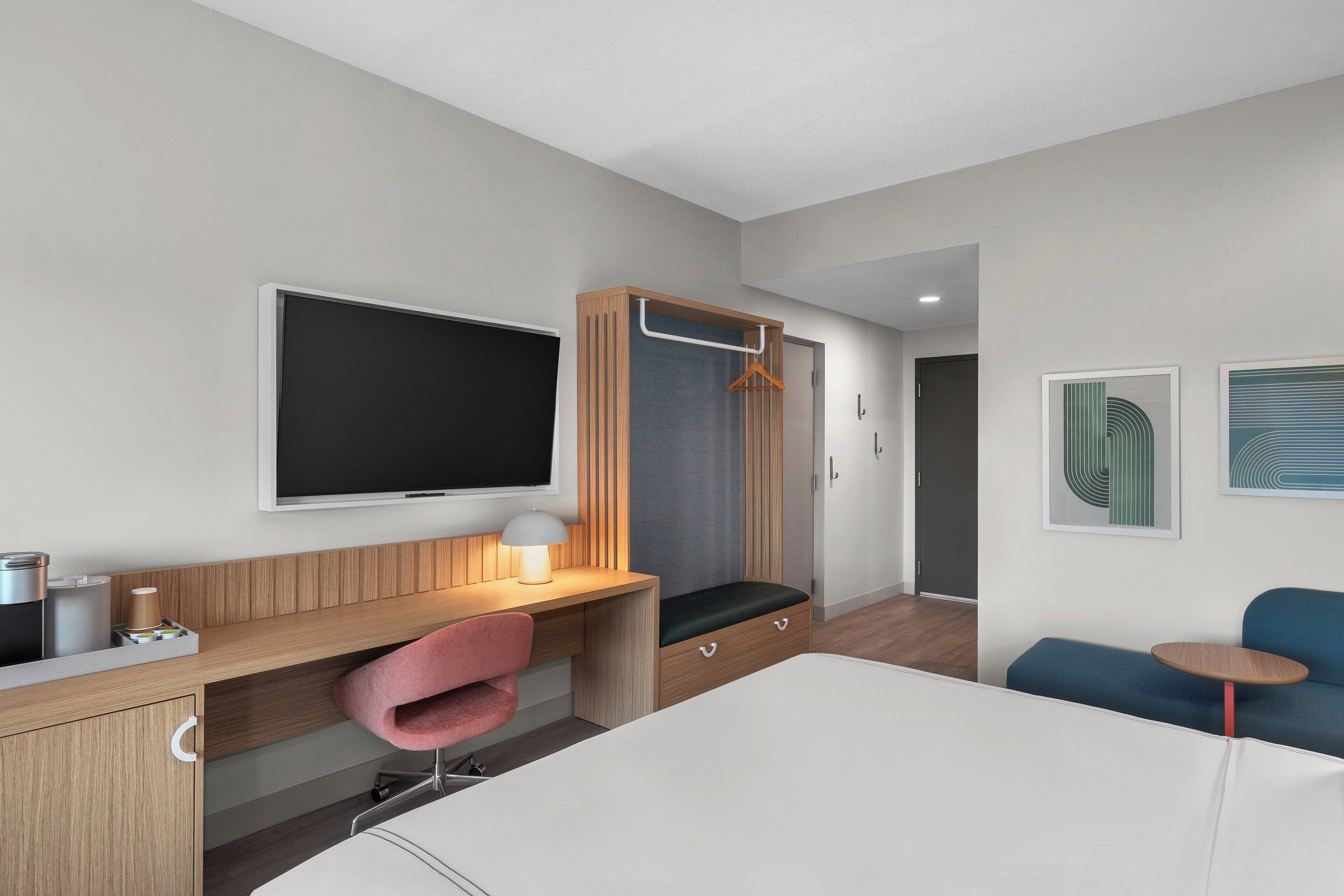 Una habitación de hotel moderna con una cama, un escritorio con una silla, un televisor, un armario y una pequeña mesa con una silla.