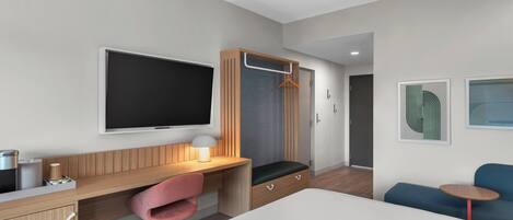 A modern hotel room with a bed, a desk with a chair, a TV, a wardrobe, and a small table with a chair.