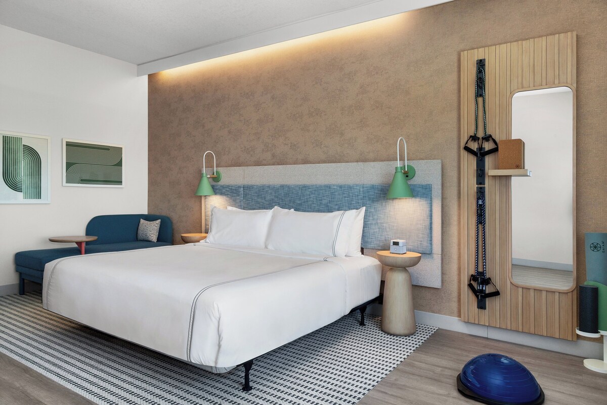 Una habitación de hotel moderna con una cama grande, dos mesitas de noche, un televisor montado en la pared, un espejo y una esterilla de yoga en el suelo.