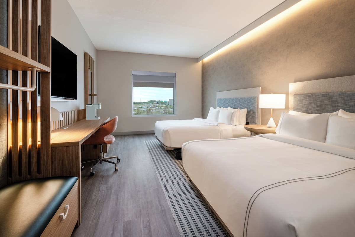 Habitación de hotel con dos camas, un escritorio, una silla, un televisor y ventana con vista a la ciudad.