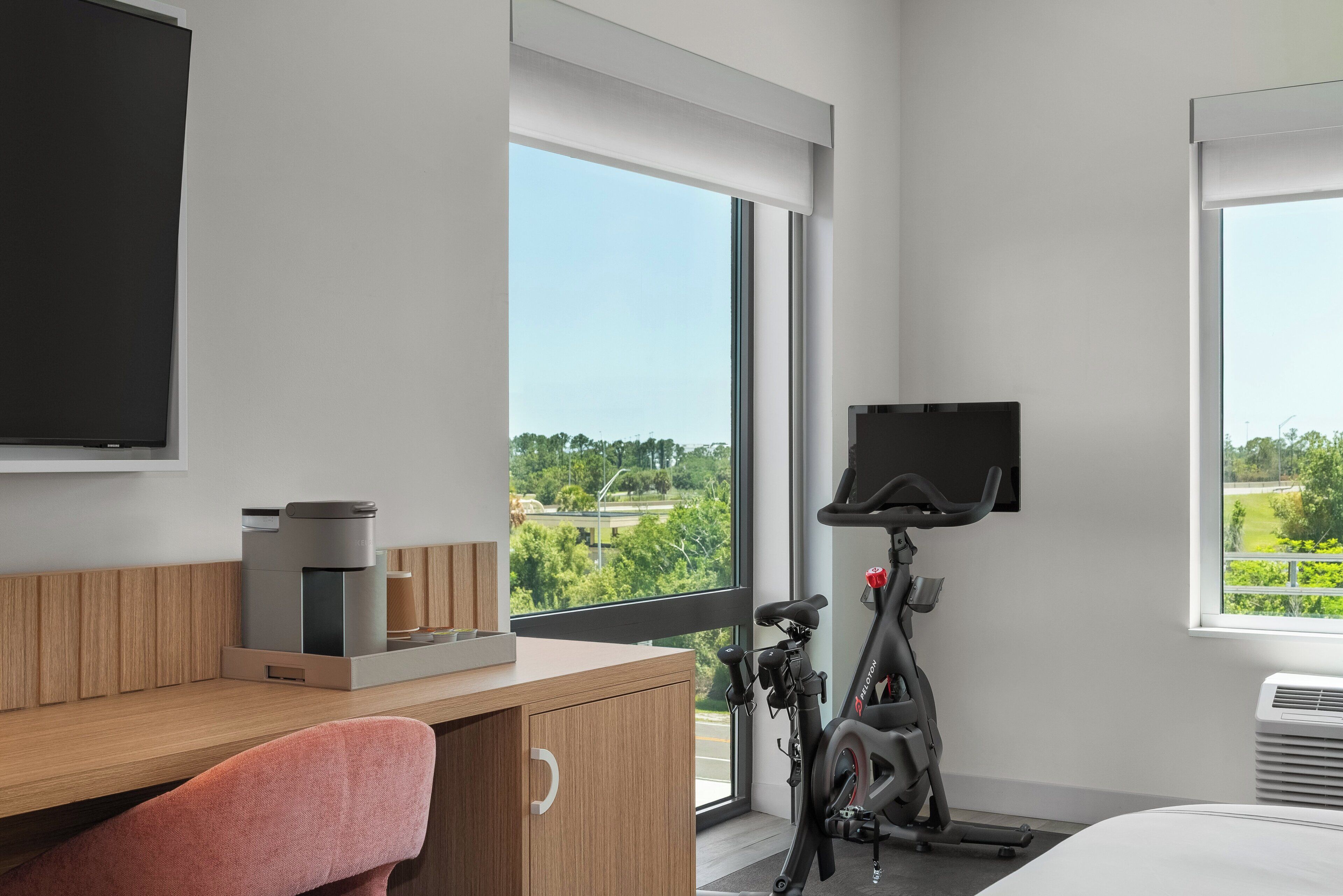 Habitación de hotel moderna con un ventanal grande, un escritorio, una silla, un televisor y una bicicleta estática.
