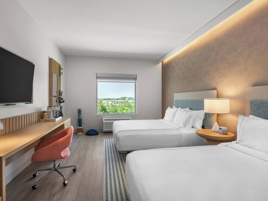 A modern hotel room with two beds, a desk with a chair, a television, and a window with a city view.