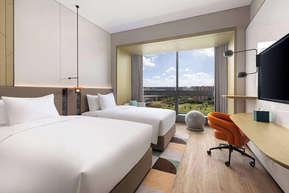 Ein Hotelzimmer mit zwei Betten, einem Schreibtisch, einem Fernseher und einem großen Fenster mit Blick auf die Stadt.