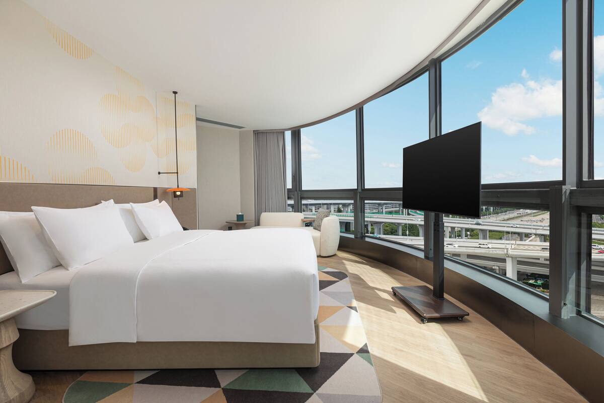 Ein modernes Hotelzimmer mit einem großen Bett, einem Fernseher und Blick auf eine Stadtlandschaft durch große Fenster.
