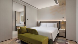 Une chambre d’hôtel moderne avec un grand lit, un canapé vert, une table de chevet avec une lampe et une salle de bain visible à travers une porte ouverte.