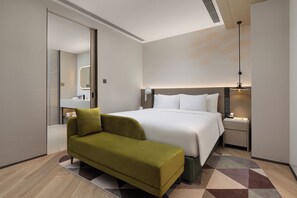 Een moderne hotelkamer met een groot bed, een groene sofa, een nachtkastje met lamp en een zichtbare badkamer door een geopende deur.