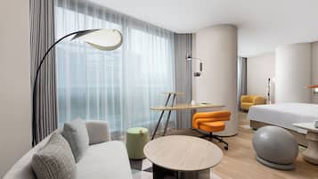 A modern hotel room with a sofa, a round table, a desk, a bed, and a large window with curtains.