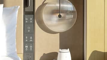 A hotel room with a bed, a bedside table with a lamp, and a control panel with various buttons.