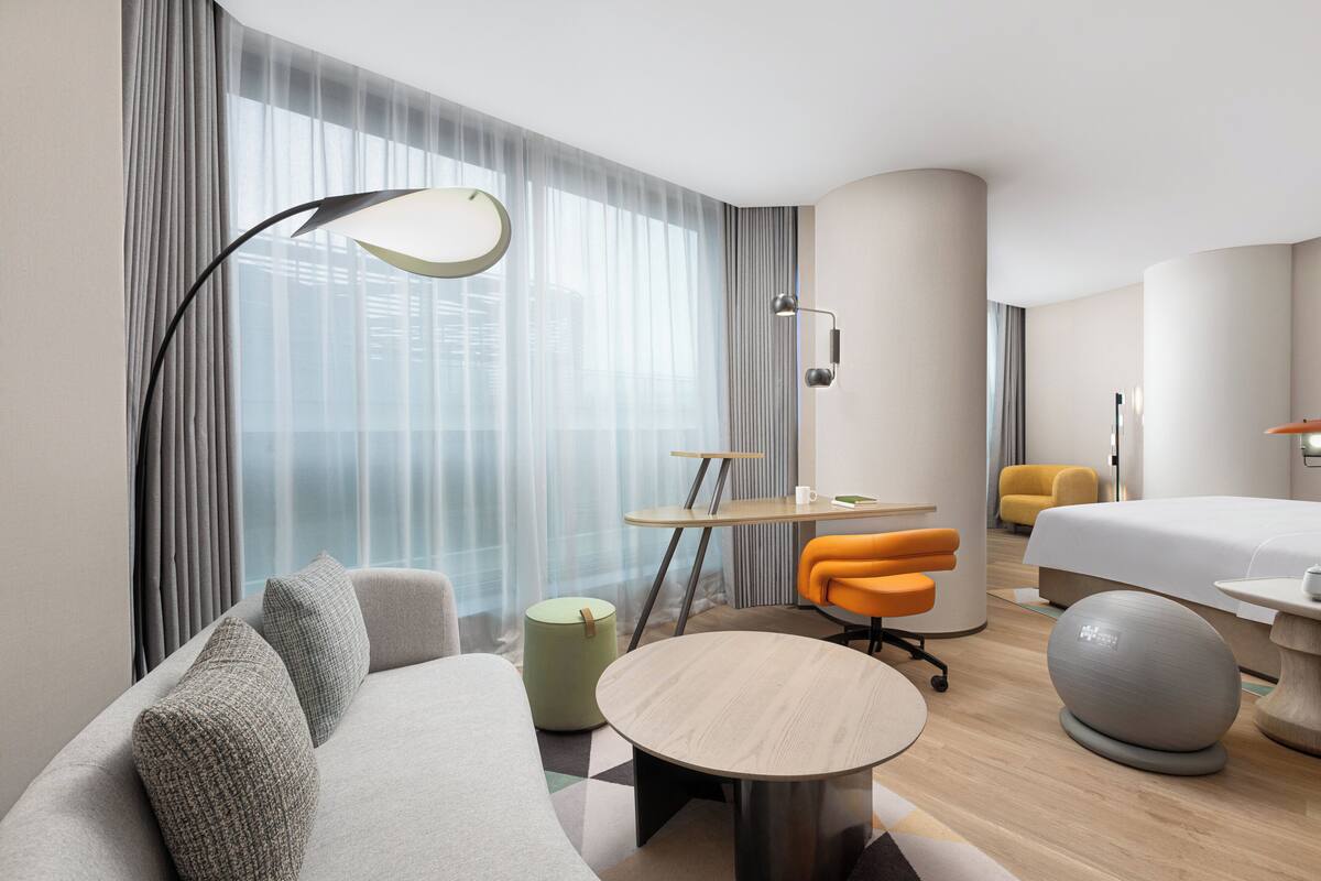 Ein modernes Hotelzimmer mit Sofa, rundem Tisch, Schreibtisch, Bett und einem großen Fenster mit Vorhängen.