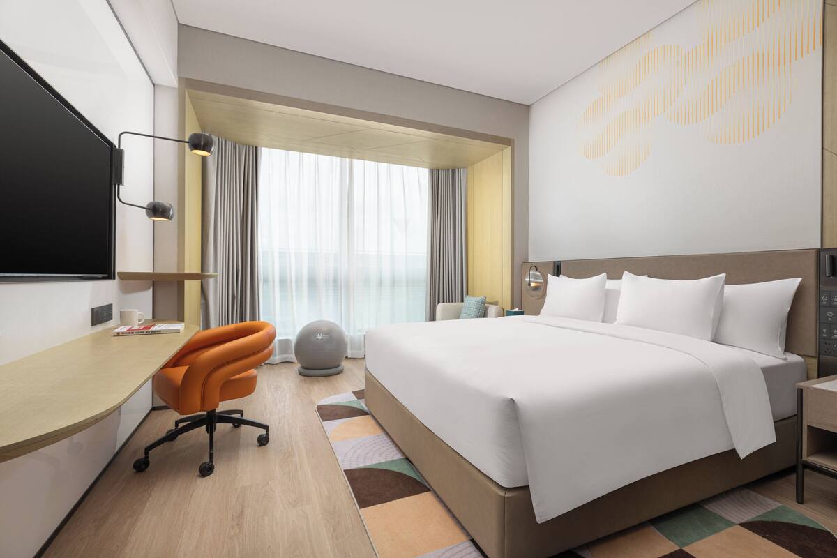 Ein Hotelzimmer mit einem großen Bett, einem Schreibtisch mit Stuhl, einem Fernseher und Blick ins Freie durch ein Fenster mit Vorhängen.