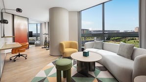 Una habitación de hotel moderna con un ventanal grande, un sofá, una mesita, una silla y un espacio para ejercitarse.