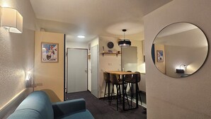 A compact hotel room with a blue sofa, a small dining table with chairs, a round mirror, and a door leading to another room. - Le balcon du soleil, Pied de pistes 2100m 5 pers, Plagne Soleil, heart of the resort (La Plagne-Tarentaise)