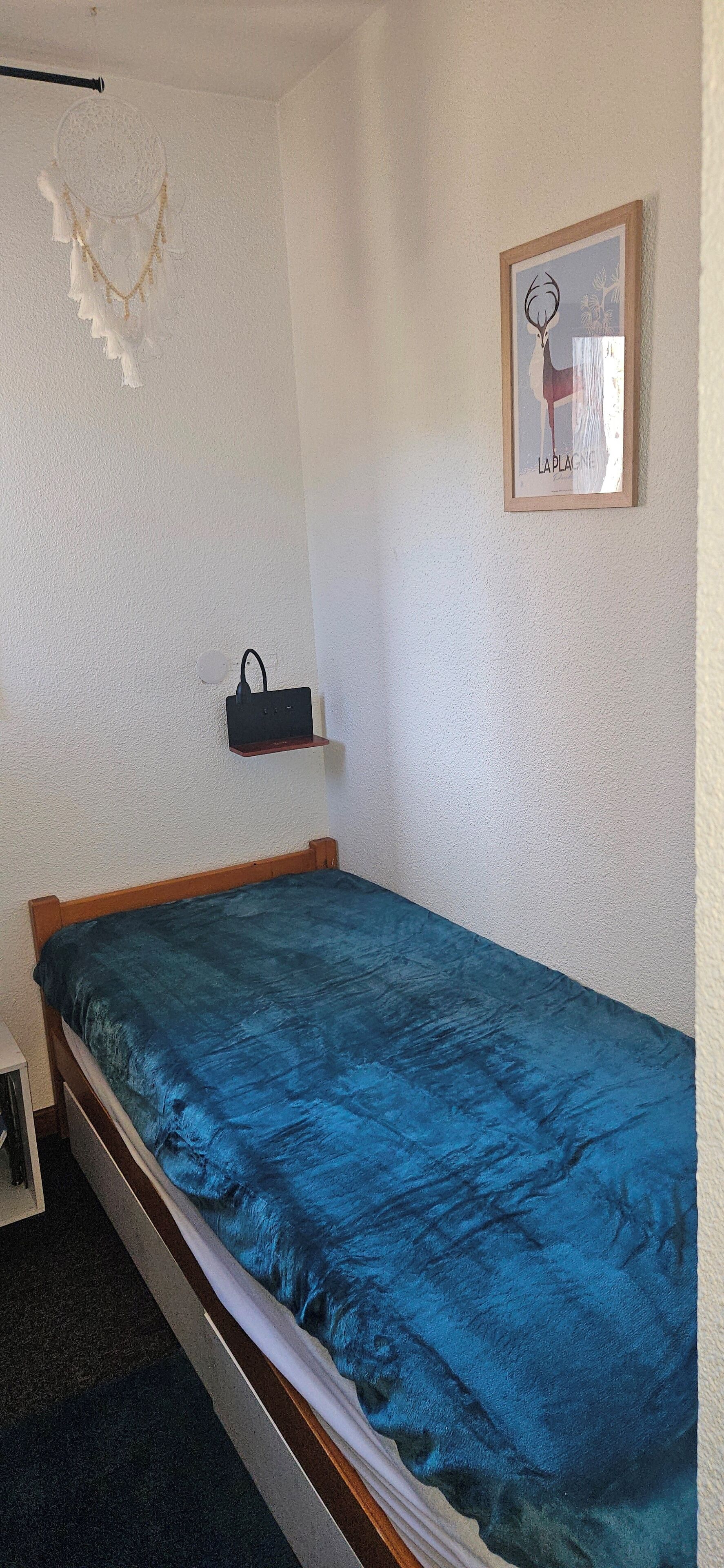 A bedroom with a single bed, a wall-mounted shelf with a bag, and a framed picture.