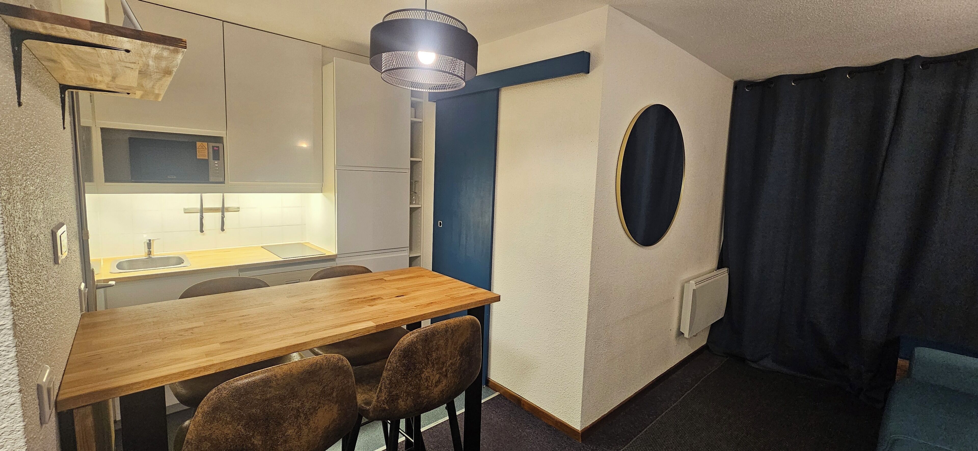 Een kleine keuken met een magnetron, spoelbak en een houten eettafel met stoelen.