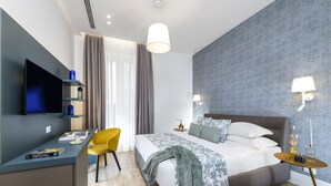 A modern hotel room with a bed, a desk, a TV, a chair, and a bedside table with lamps. - Domus Augustea - Cesare (Rome)