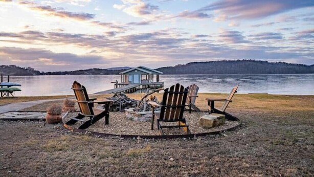 Una cabaña junto al lago con un muelle, rodeada de sillas Adirondack, un hoyo para fogatas y un cobertizo pequeño.