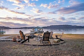 A lakeside cabin with a dock, surrounded by Adirondack chairs, a fire pit, and a small shed.