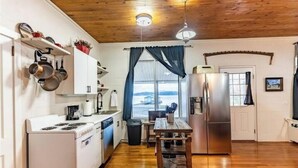 Cocina compacta con gabinetes blancos, estufa y refrigerador. Hay una ventana con cortinas, techo de madera y vista a un edificio.