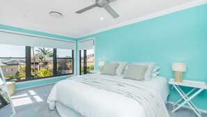 Ein Schlafzimmer mit einem großen Bett, einem Deckenventilator und Blick auf eine Wohngegend durch ein großes Fenster.