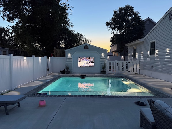 Una piscina con una pantalla de televisión, una valla blanca y una casa al fondo.