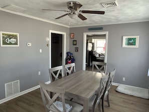 Un comedor con una mesa y sillas de madera, un ventilador de techo y una cocina visible a través de un vano abierto.