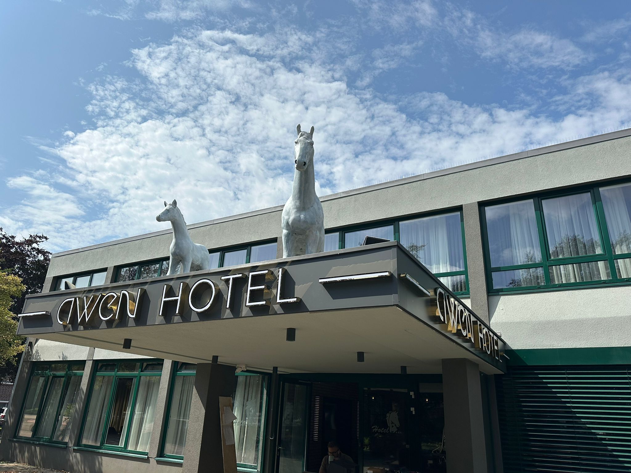 Ein Hotel mit moderner Fassade, zwei Pferdestatuen auf dem Dach und blauem Himmel im Hintergrund.
