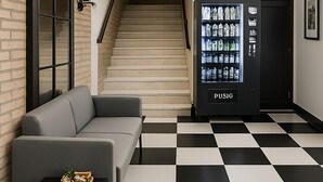 A lobby with a checkered floor, a vending machine, a staircase, and a grey sofa. - Awen Hotel Vechta (Vechta)