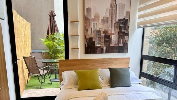 Quarto moderno com uma cama grande, uma pintura de paisagem urbana e vista para um pátio com mesa e cadeiras.