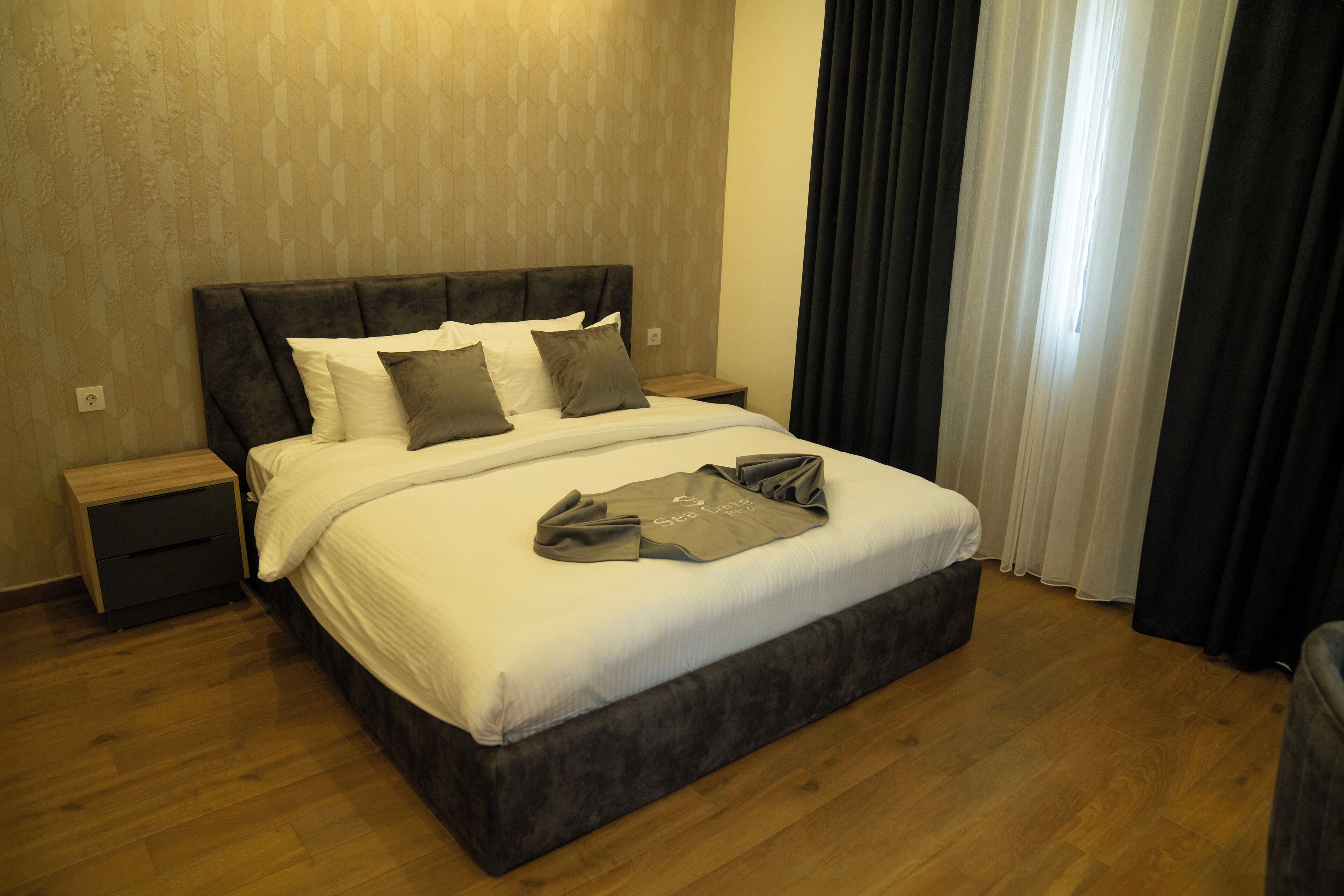 Una cama bien tendida con un cabecero oscuro, una mesita de noche de madera y una pared de color claro con un diseño geométrico.