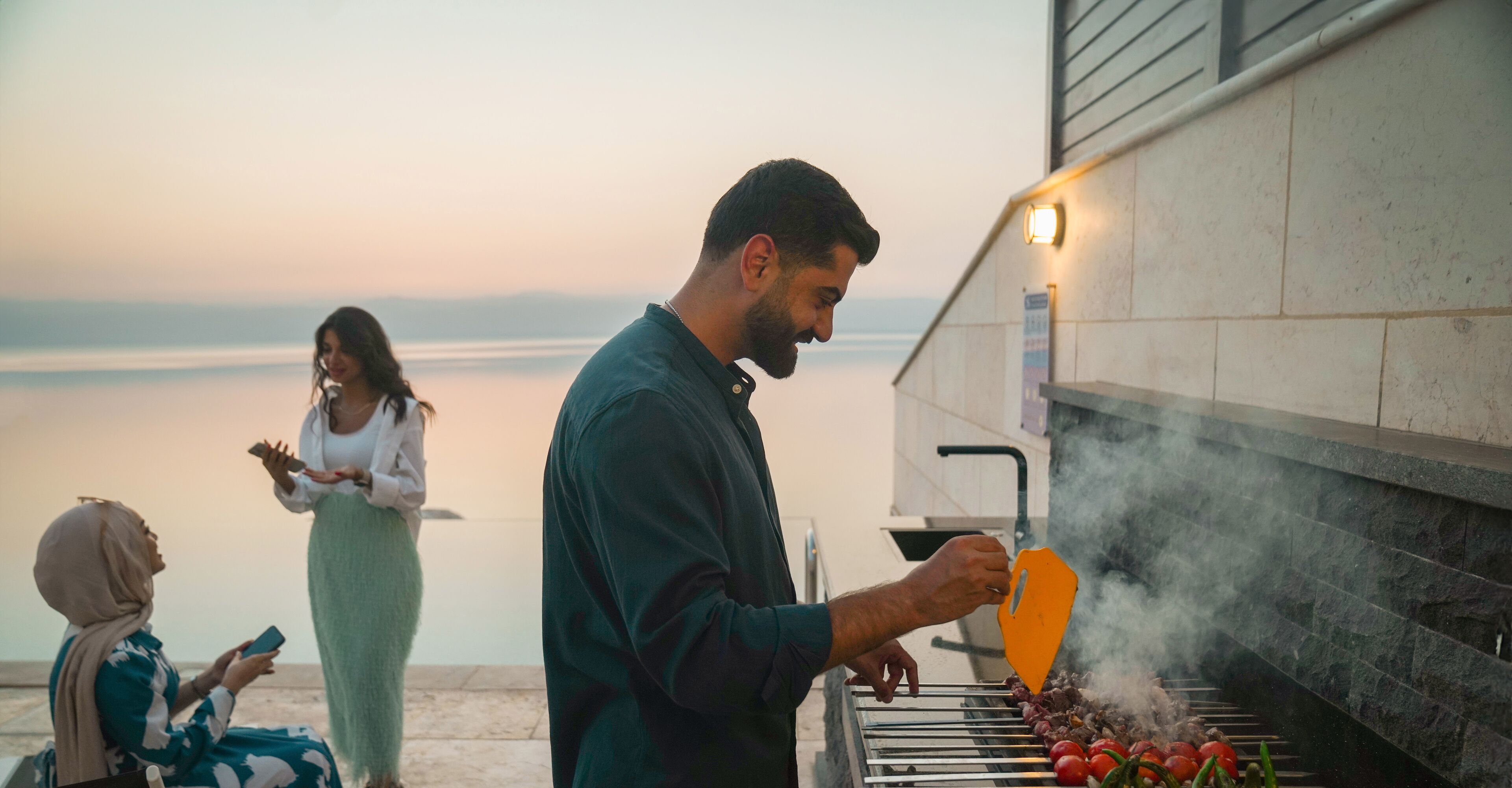Ein Mann grillt Essen auf einem Grill im Freien, im Hintergrund ist das Meer zu sehen.