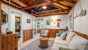 A cozy room with a wooden ceiling, a white sofa, a wicker ottoman, and a view of the outdoors. - Lakefront Cottage Retreat: 3BR w/ Fire Pit, BBQ & Dock (Plymouth)