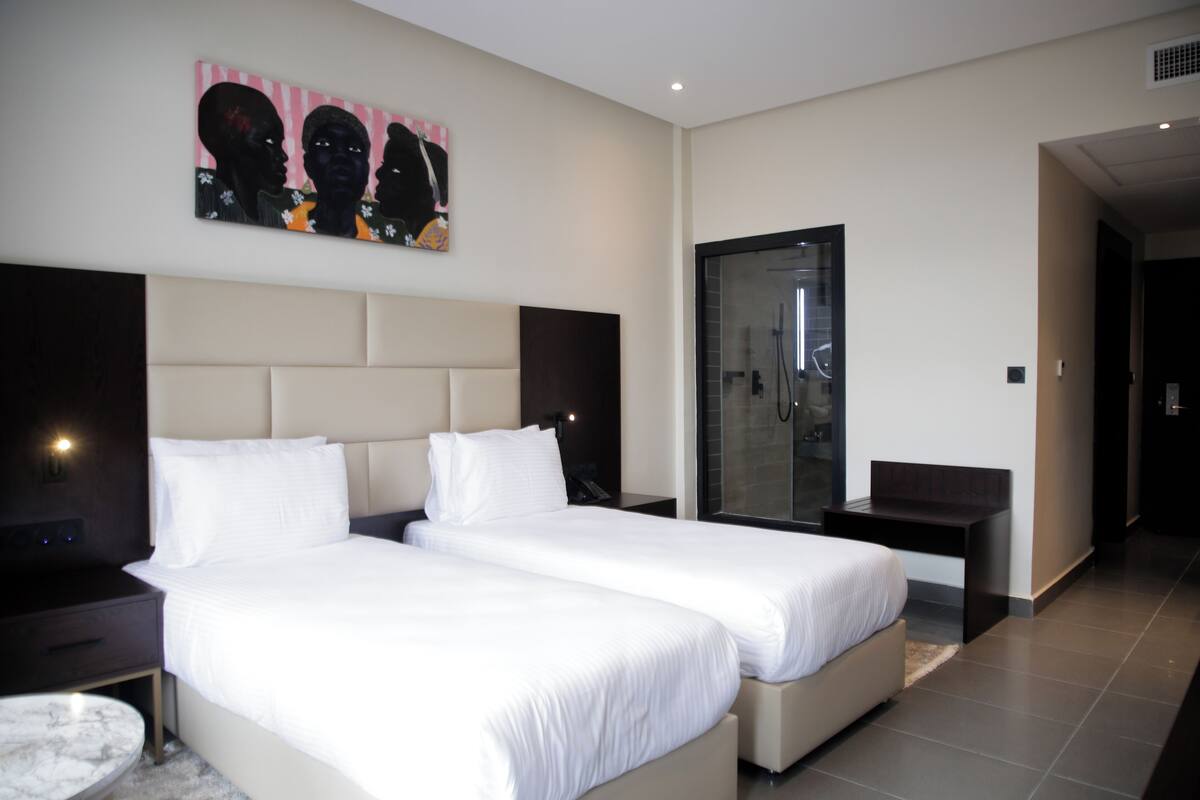 A modern hotel room with two beds, a large artwork on the wall, a glass-enclosed shower, and a marble-tiled floor.