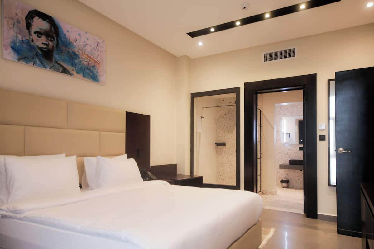 A modern hotel room with a large bed, a painting on the wall, a shower area, and a door leading to a bathroom.