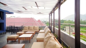 Eine Dachterrasse mit Außenmöbeln, darunter Sofas und Tische, und Blick auf ein Gebäude mit rotem Dach sowie grüne Elemente im Hintergrund.