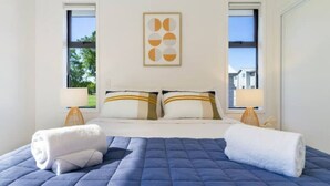 Un lit bien fait, recouvert d’une couette bleue, avec deux oreillers à motifs rayés et deux serviettes blanches. Au-dessus du lit, on aperçoit une œuvre d’art encadrée, et une fenêtre se trouve sur le côté gauche.