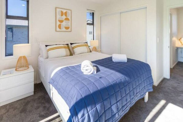 Una cama bien tendida con colcha azul, almohadas blancas y una toalla enrollada encima. Hay una lámpara de noche a la izquierda y un cuadro enmarcado en la pared sobre la cama.