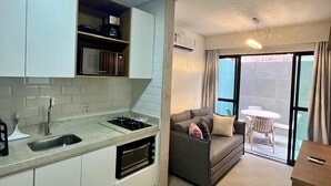A modern kitchen with white cabinets, a built-in oven, and a sink. There is a grey sofa and a small table with chairs in the living area. - Green Porto 02 (Ipojuca)