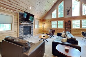 A log cabin interior with a fireplace, leather sofas, a flat-screen TV, and large windows. - The Pour House by Taylor Made Deep Creek (McHenry)