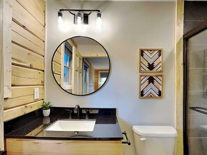 A bathroom with a wooden vanity, a round mirror, a sink, a toilet, and two decorative wall pieces. - The Pour House by Taylor Made Deep Creek (McHenry)