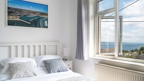 A bedroom with a bed, a nightstand, a window with a view of a coastal town, and a painting on the wall.