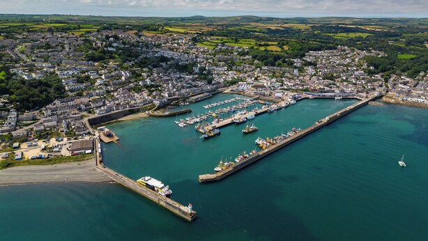 An aerial view of a coastal town with a marina, residential areas, and surrounding green fields.