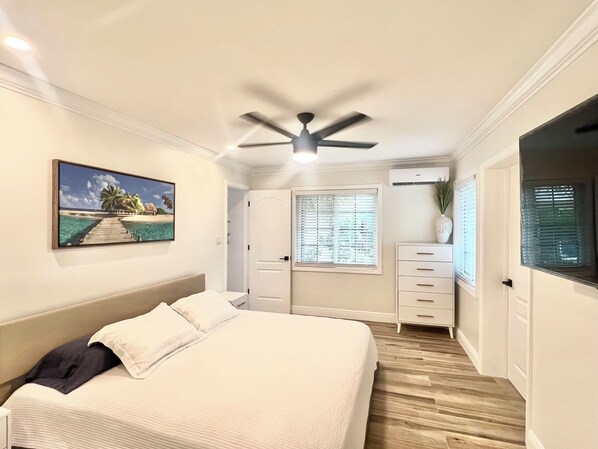 Une chambre à coucher avec un lit, un ventilateur de plafond, un tableau, une commode et une fenêtre avec des stores.