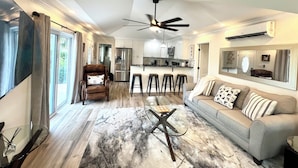 A modern living room with a sofa, a coffee table, and a TV. There is a kitchen area with a refrigerator and a dining table with bar stools.