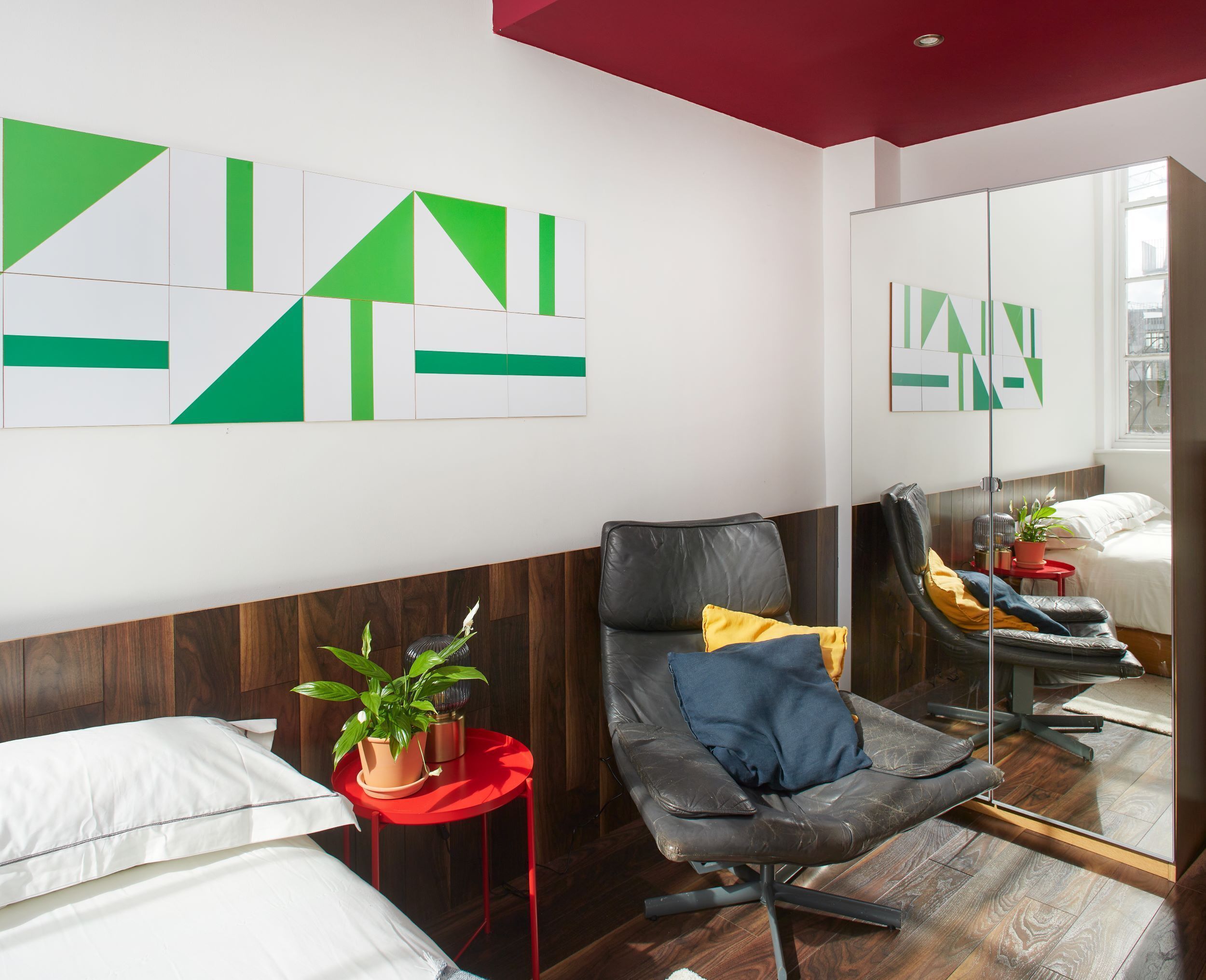 A modern hotel room with a wooden floor, a black leather chair, a red side table with a plant, and a large mirror reflecting the room.