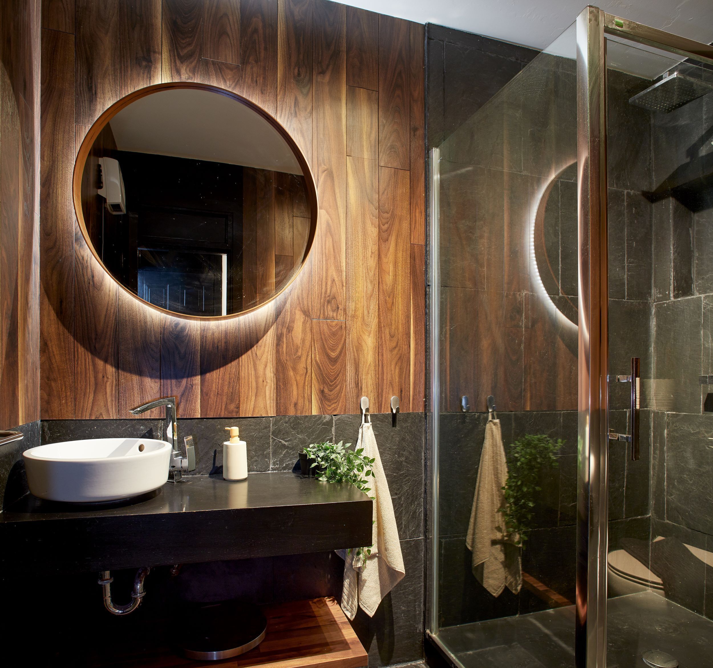 Un baño moderno con tocador de madera, espejo redondo, lavamanos blanco y cabina de ducha de cristal.