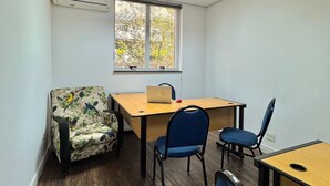 A small, well-lit room with a wooden desk, two chairs, a patterned armchair, a window with a view of trees, and an air conditioning unit. - Get a Flat 601 Ibirapuera -Jardins (São Paulo)